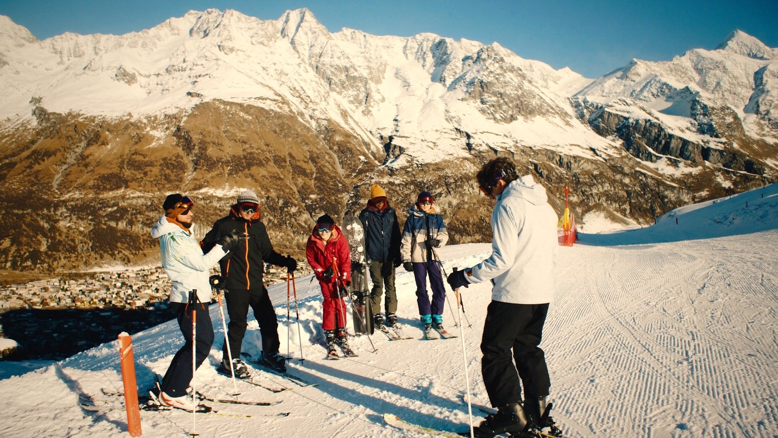 A group of skiers laughing together at the top of a sunny groomed run above an alpine village