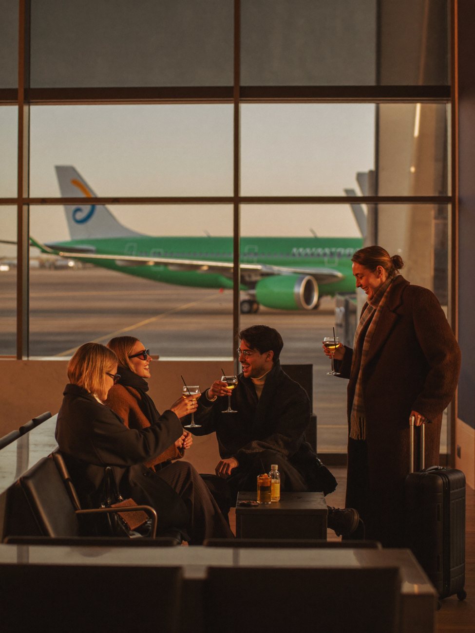 A group toasting cocktails in an airport lounge at golden hour, a jet beyond the glass